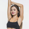 Woman with long hair wearing a black sports bra, arms raised overhead in a stretch against a white background.