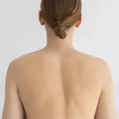 Back view of a woman with her hair tied into a low bun secured with a hair tie, bare shoulders visible against a neutral backdrop.