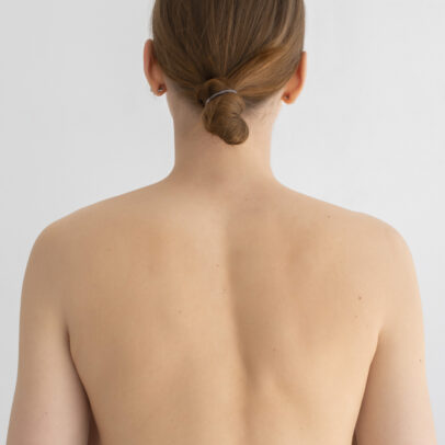 Back view of a person with light brown hair in a small bun secured by a gray hair tie; bare shoulders and upper back visible.