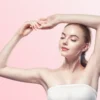 Woman with eyes closed, arms raised above her head in a relaxed pose against a pink gradient background, wearing a white strapless top.