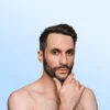Close-up portrait of a shirtless man with a beard, touching his chin and looking at the camera against a soft blue gradient background.