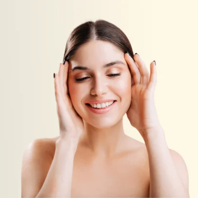 Smiling woman cupping her cheeks with both hands, eyes closed, showcasing clear skin against a pale background.