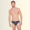 Male model wearing dark gray briefs, standing facing the camera against a pale gradient background.