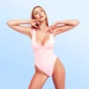 Woman in a pink one-piece swimsuit poses with arms bent and eyes closed against a pale blue gradient background, exuding confidence.