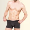 Man standing shirtless, wearing black boxer briefs and adjusting waistband against a light beige gradient background.