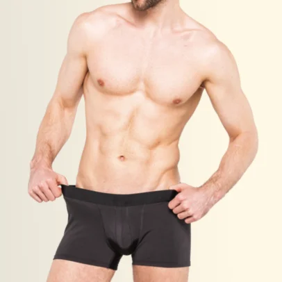 Man standing shirtless, wearing black boxer briefs and adjusting waistband against a light beige gradient background.