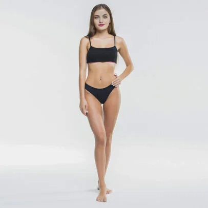 Full-body portrait of a woman in a black bikini standing barefoot in a studio, hand on her hip.