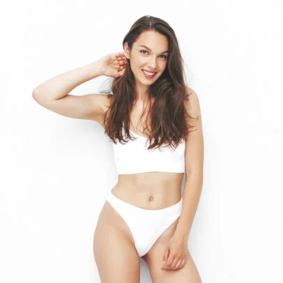 Smiling woman in a white bra and high-cut panties posing with one hand behind her head against a white background.