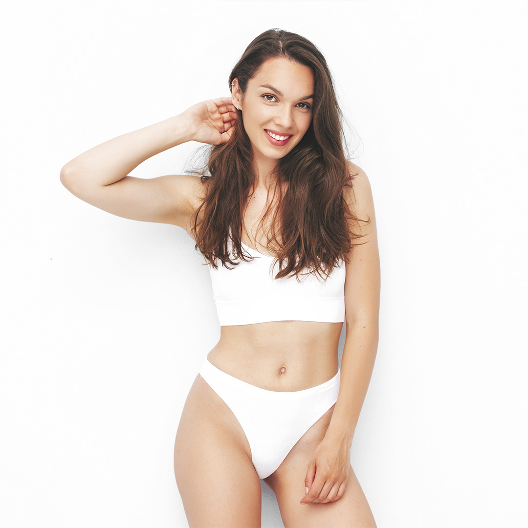 Smiling woman in a white bra and high-cut panties posing with one hand behind her head against a white background.