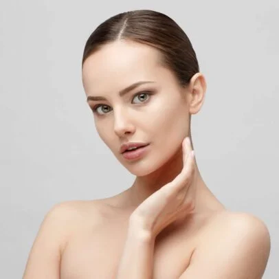 Close-up portrait of a young woman with smooth skin touching her jawline against a light gray background, looking at the camera.