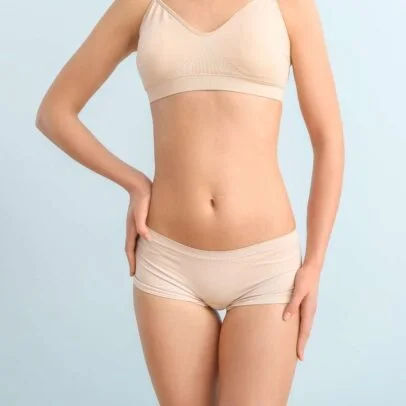Midsection of a woman wearing beige bra and underwear, standing with one hand on her hip against a pale blue background.