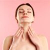 Woman with eyes closed performing a neck/throat massage, hands at the front of her neck against a pink background (self-care moment).