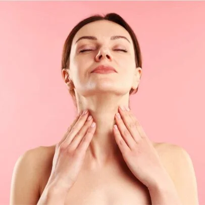 Woman with eyes closed performing a neck/throat massage, hands at the front of her neck against a pink background (self-care moment).
