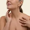 Close-up of a woman with bare shoulders, one hand at her jaw and the other on her chest.