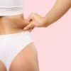 Person pinching a roll of fat at the waist, wearing white underwear against a pink background.