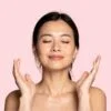 Woman with eyes closed, smiling softly, hands raised in a gentle meditation pose against a pink background.