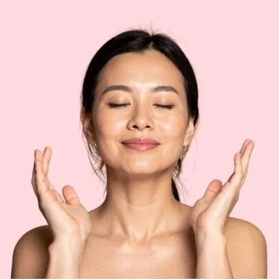 Woman with eyes closed, smiling softly, hands raised in a gentle meditation pose against a pink background.