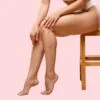 Lower body of a woman sitting on a wooden stool with legs crossed, against a pink background.