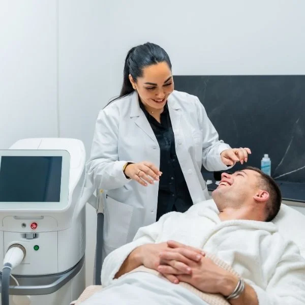Medical professional in a white coat performing a treatment on a man lying in a clinic bed next to a medical device with a touchscreen monitor.